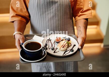Horizontale Aufnahme aus einem hohen Winkel der nicht erkennbaren Kellnerin, die im Café arbeitet und Kaffee und Dessert zum Kunden bringt Stockfoto