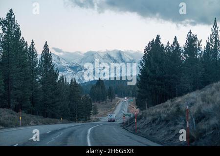 Verkehrsstraße entlang des dichten Waldes mit verschneiten Bergen im Hintergrund an einem düsteren Tag Stockfoto