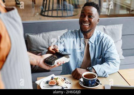Fröhlicher junger schwarzer Mann, der am Tisch in einem gemütlichen Café sitzt und mit seinem Smartphone für das Mittagessen bezahlt Stockfoto