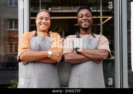 Horizontales mittleres Porträt eines glücklichen jungen schwarzen Mannes und einer glücklichen Frau, die Schürzen tragen und die Arme am Eingang des Cafés mit einem Lächeln auf die Kamera stehen Stockfoto