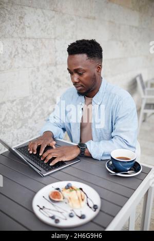 Porträt eines jungen schwarzen Mannes in stilvollem legerem Outfit, der im Freien am Kaffeetisch sitzt und mit Freunden auf dem Laptop plaudert Stockfoto