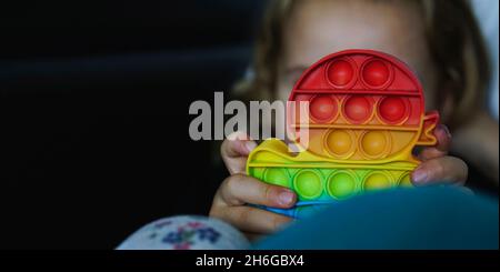 Kleines Kind spielt mit Regenbogen Pop-it Fidget Gadget. Beliebtes Anti-Stress-Spielzeug aus Silikon. Jugendkultur und Konzept der psychischen Gesundheit. Modisches, flexibles Spielzeug Stockfoto