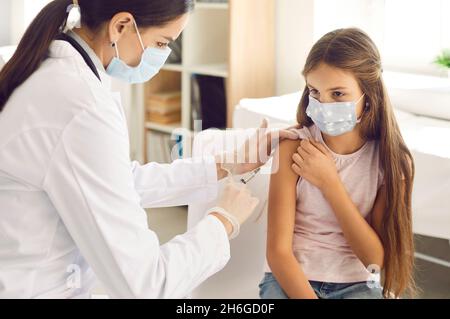 Nachdenkliches kleines Mädchen in Gesichtsmaske wird vom Arzt oder der Krankenschwester in der Klinik gegen Coronavirus geimpft. Stockfoto