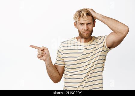 Porträt eines verärgerten blonden Typen, der nach links zeigt, die Hand auf den Haarschnitt hält und unzufrieden grinst, weißer Hintergrund Stockfoto