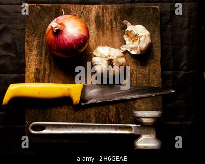 Draufsicht auf eine Zwiebel- und Knoblauchzwiebeln auf einem Schneidebrett mit einem Messer und einem Hammer darauf Stockfoto