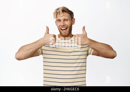 Lächelnder, gutaussehender Mann, der Daumen hoch zeigt und zwinkert, sagt ja, Lob und Kompliment und steht im T-Shirt auf weißem Hintergrund Stockfoto
