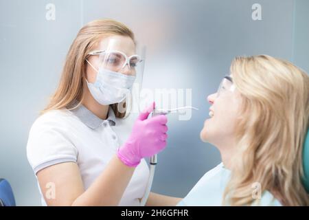 Zahnärztin mit medizinischer Gesichtsmaske und plastischem Gesichtsschutz, die während einer Coronavirus-Pandemie mit weiblicher Patientin zusammenarbeitet Stockfoto