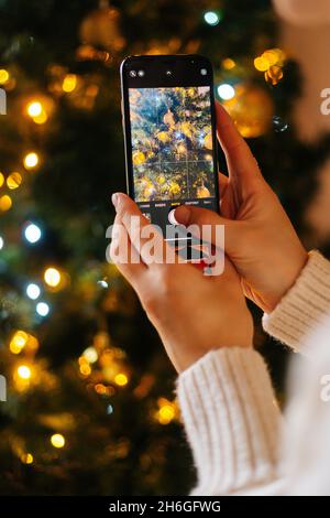 Vertikale Nahaufnahme einer nicht erkennbaren jungen Frau, die zu Hause Fotos macht oder Videos von der Dekoration des Weihnachtsbaums und der Feier von Bokeh-Lichtern macht Stockfoto
