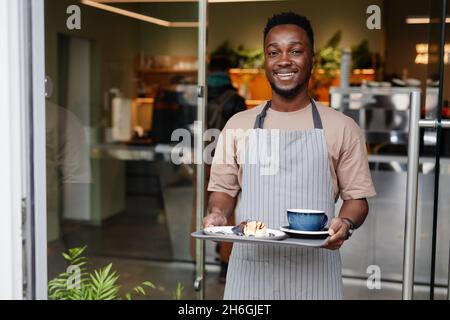 Horizontale mittlere Aufnahme eines gutaussehenden jungen Mannes, der in einem Café mit Tablett und Kaffee und Dessert auf dem Tablett arbeitet und die Kamera anlächelt Stockfoto