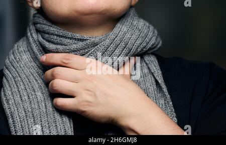 Ein grauer Schal ist um den Hals gewickelt, und eine Hand hält den Hals. Erkältung und Halsschmerzen Stockfoto