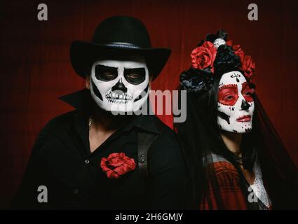 Schöne Frau mit einem Zuckerschädel Make-up mit einem Blumenkranz auf ihrem Kopf und einem Skelett Mann in einem schwarzen Hut. Paar auf dunkelrotem Hintergrund Stockfoto