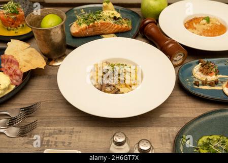 Internationale Gerichte auf dunklem Holztisch. Blisines, Pasta mit Parmesan, gegrillter norwegischer Lachs, Thunfisch-Tartare, Eier benedict Stockfoto