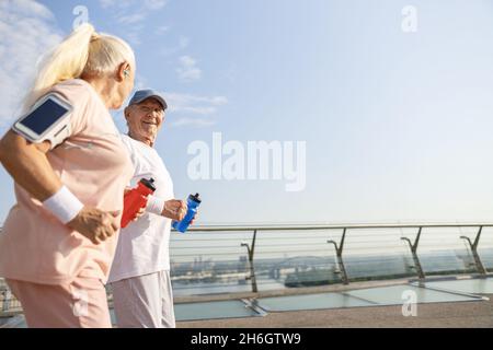 Fröhlicher älterer Mann und Frau mit Wasserflaschen laufen entlang der Fußgängerbrücke der Stadt Stockfoto
