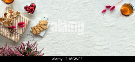 Panorama-Herbstbanner. Herbstdekorationen - Preiselbeere, Kekse. Tee und trockene Eichenblätter. Flach liegend, Draufsicht auf unfarbigem Textil. Herbsthintergrund Stockfoto