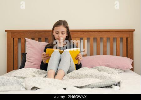 Mädchen im jungen Schulalter, das im Schlafzimmer auf dem Bett sitzt und aus dem gelben Lehrbuch liest. Schulaufgaben. Stockfoto