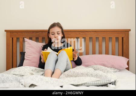 Mädchen im jungen Schulalter, das im Schlafzimmer auf dem Bett sitzt und aus dem gelben Lehrbuch liest. Schulaufgaben. Stockfoto