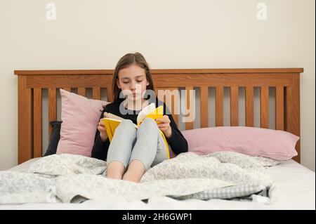 Mädchen im jungen Schulalter, das im Schlafzimmer auf dem Bett sitzt und aus dem gelben Lehrbuch liest. Schulaufgaben. Stockfoto