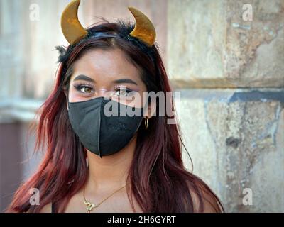 Hübsches mexikanisches Mädchen mit gelben Teufelshörnern Stirnband trägt während der globalen Coronavirus-Pandemie eine schwarze Gesichtsmaske und schaut auf den Zuschauer. Stockfoto