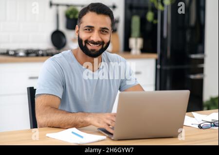 Porträt eines gutaussehenden indischen bärtigen Mannes, Freiberuflers, Designers oder IT-Spezialisten, der von zu Hause aus mit einem Laptop arbeitet, während er am Tisch in der Küche sitzt, direkt in die Kamera blickt und freundlich lächelt Stockfoto