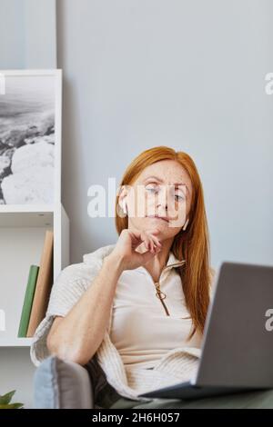 Vertikales Porträt einer erwachsenen rothaarigen Frau, die in einem Online-Meeting zu Hause einen Laptop verwendet Stockfoto