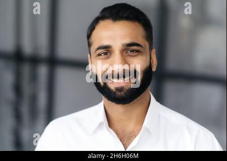 Nahaufnahme eines erfolgreichen, intelligenten bärtigen indischen Geschäftsmannes, Managers oder IT-Spezialisten, der ein weißes Hemd trägt und mit einem freundlichen Lächeln auf die Kamera blickt Stockfoto