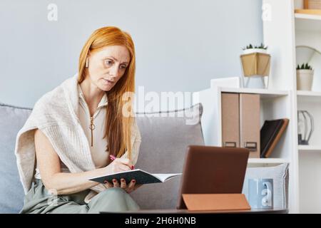 Porträt einer erwachsenen rothaarigen Frau, die zu Hause online studiert und Notizen macht, Platz kopiert Stockfoto