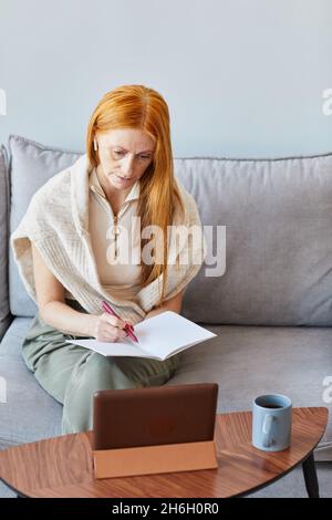 Vertikales Porträt einer erwachsenen rothaarigen Frau, die zu Hause online studiert und sich Notizen gemacht hat Stockfoto
