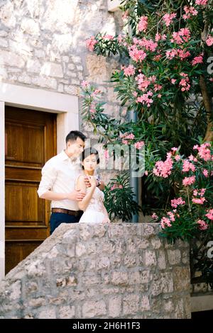 Sibenik, Kroatien - 05.06.17: Bräutigam umarmt die Braut in einem weißen Spitzenkleid von hinten an den Schultern, während er auf der Veranda eines alten Hauses in der Nähe eines Stockfoto