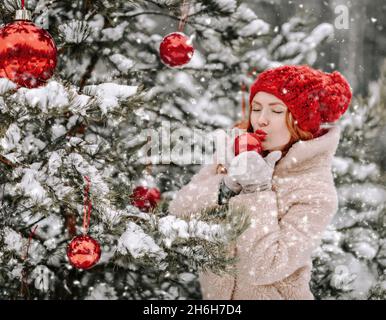 Junge rothaarige Frau im Pelzmantel steht draußen am geschmückten Weihnachtsbaum und küsst einen roten Ball unter Schnee Stockfoto