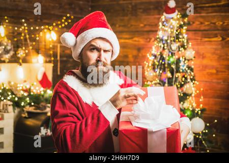 Hipster in rotem Weihnachtsmann Hut hält präsent. Styling Santa Mann mit einem langen Bart posiert auf dem Weihnachts-Holzhintergrund. Stockfoto