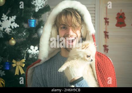 Neujahr des Hundes, erstaunt Mann halten Welpen. Weihnachtsmann mit Haustier am Baum. Weihnachtsmann mit glücklichem Gesicht und Haustier, pommerscher Spitz. Stockfoto