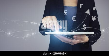 Erfolgreiches internationales Finanzinvestitionskonzept. Weißes Tablet in Geschäftsfrau Hand mit digitalen Hologramm Weltwährungen und Pfeil nach oben auf grau Stockfoto