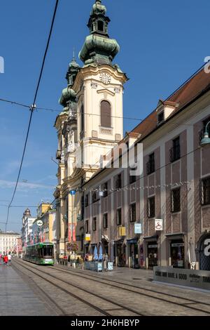 Linz, Österreich - 26. September 2021: Altstadt von Linz mit bunten Straßenbahnen, die vor einer historischen Kirche vorbeifahren Stockfoto