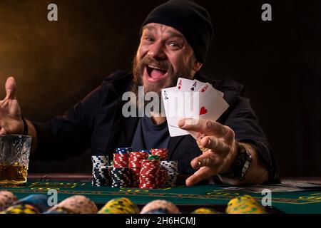 Bärtiger Mann mit Zigarre und Glas sitzt am Pokertisch und schreit isoliert auf Schwarz Stockfoto