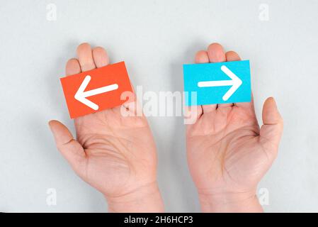 Ein blaues und ein rotes Blatt Papier, das in den Händen hält, Pfeile, die verschiedene Richtungen zeigen, eine Entscheidung treffen, eine Chance wählen Stockfoto