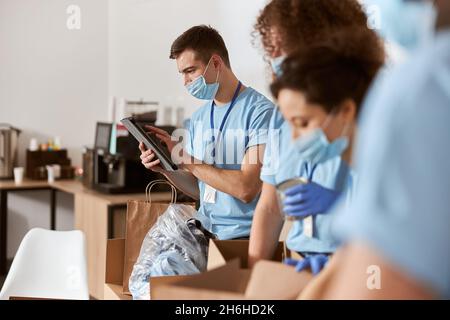 Eine Gruppe von fleißigen Freiwilligen, die blaue Uniform, Schutzmasken und Handschuhe tragen und dabei die gespendeten Lebensmittel berechnen und sortieren, während sie in gemeinnützigen Einrichtungen arbeiten Stockfoto