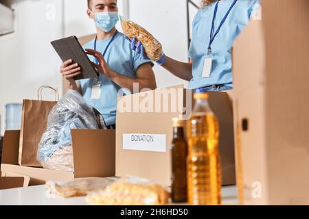 Nahaufnahme von Freiwilligen mit blauer Uniform, Schutzmasken und Handschuhen, die während der Arbeit in Kartons gespendete Lebensmittel sortieren Stockfoto