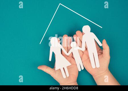 Eine Familie, Mutter, Vater und Kind in Silhouette, Papier ausgeschnitten, in den Händen haltend, Dach eines Hauses, petrofarbener Hintergrund Stockfoto