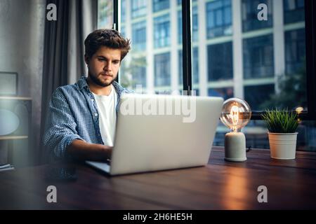 Junger Mann, der mit einem Laptop arbeitet. Freiberuflicher Mitarbeiter, der über einen Computer mit dem Internet verbunden ist. Blogger oder Journalist, der einen neuen Artikel schreibt. Stockfoto