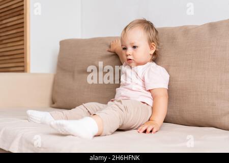 Kleines Mädchen, das auf dem Sofa sitzt und mit Interesse wegschaut. Blonde blauäugige Baby in hellen Kleidern Stockfoto