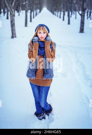 Liebenswert Winter Mädchen tragen Pelzweste, Jeans und Pullover Spaß im Winter Park. Schöne kaukasische junge Frau in einem Schal gewickelt Blick auf die Kamera. Modelmodell. Vertikale Aufnahme Stockfoto