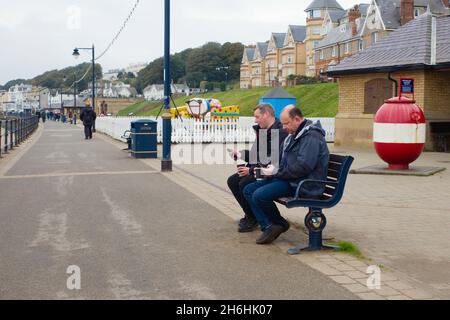 Zwei Männer saßen auf einer Bank am Meer, tranken Kaffee und schauten sich ihre mobilen Geräte in Filey an. Stockfoto