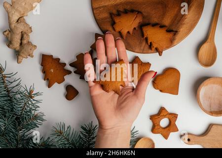 Weibliche Hand hält Kekse über Weihnachts-Lebkuchen in Form von Sternen und Weihnachtsbäumen auf weißem Tisch mit Zweigen der Weihnachtstanne. Oben vi Stockfoto