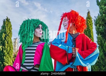 Positive männliche und weibliche Harlekine mit bemalten Gesichtern in bunten Kostümen und Perücken, lächelnd und schauend einander während des Festivals in Ark an sonnigen Tagen an Stockfoto