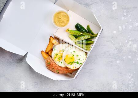 Spiegeleier mit Wurst, Kartoffelkeilen und eingelegter Gurke in Papppackungen zum Mitnehmen Stockfoto