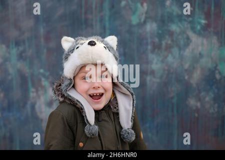 Porträt eines überraschten Kindes in einem Wolfhut und lächelt sanft auf grauem Hintergrund, Blick auf die Kamera, im Freien. Platz für Text. Hochwertige Fotos Stockfoto