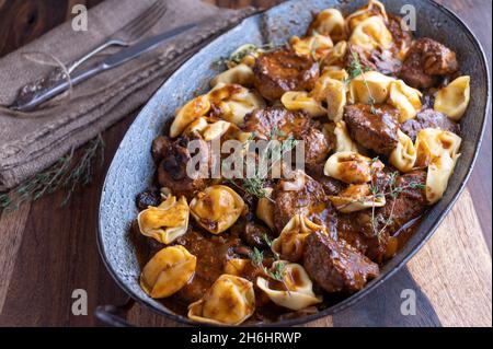 Geschmortes Schweinefilet mit einer köstlichen Pilzsauce, serviert mit Tortellini in einer rustikalen Bratpfanne auf einem Holztisch Stockfoto