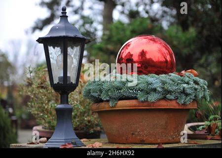 Weihnachtsdekoration im Garten. Ein flacher Terrakotta-Topf mit einem Tannenkranz und einer roten Glaskugel an einer Eingangsbeleuchtung Stockfoto