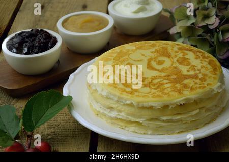 Russische Pfannkuchen crepes im Stapel und auf der Pfanne mit Apfelmus und saurer Creme auf rustikalem Holzhintergrund. Stapel von russischen Pfannkuchen Blini auf einem hölzernen Stockfoto