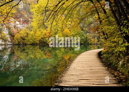 Eine rustikale Holzpromenade, die entlang des Ufers eines malerischen Bergsees mit Bäumen und Laub in intensiven Herbstfarben führt Stockfoto